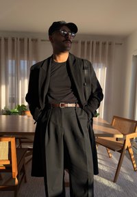 Μαύρο μπουφάν πάνω από μαύρο πουκάμισο, ψηλόμεσα μαύρα παντελόνια με πτυχές, καφέ ζώνη, μαύρο καπέλο και γυαλιά. Το φυσικό φως αναδεικνύει τις υφές.