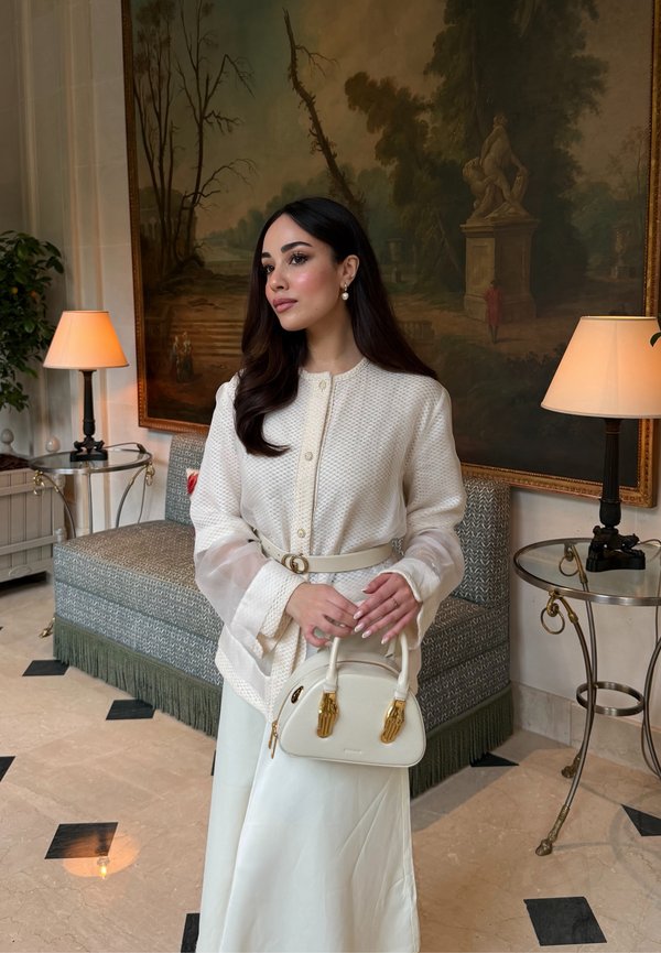 Donna con lunghi capelli scuri che indossa giacca e gonna bianche testurizzate, tiene una borsa bianca, in piedi in una stanza elegante con un quadro e lampade.