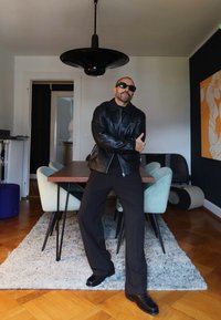 Svart skinnjakke og mørke bukser båret av en person som står i et moderne interiør, med en svart pendellampe og et trebord til spising.