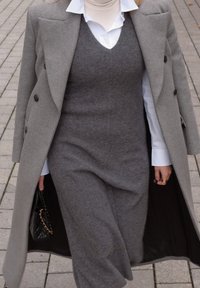 Šedý vlnený kabát cez šedé vlnené šaty s bielym golierom košele. Šaty majú priliehavý strih a drží sa čierna kabelka s reťazovými detailmi.