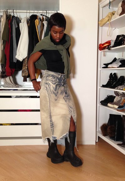 Mujer con una falda vaquera desgastada, botas negras y una camiseta negra con un suéter verde atado alrededor de los hombros, de pie en un armario.