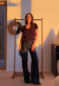 Femme portant un haut violet foncé et un jean à jambes larges, debout à l'intérieur près d'un support en bois avec une couronne, éclairée par une lumière solaire chaude.