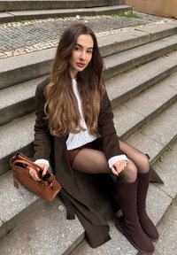 Brauner langer Mantel über einem weißen Pullover mit Zopfmuster, braune Leder-Shorts und kniehohe braune Stiefel. Eine braune Handtasche mit zwei Griffen liegt daneben.