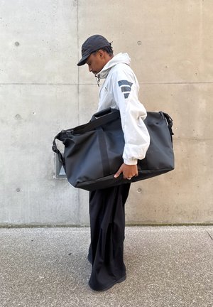 Hombre con chaqueta blanca y gorra negra llevando una gran bolsa deportiva negra, de pie frente a una pared de concreto.