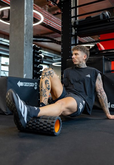 Hombre con tatuajes usando un rodillo de espuma en la pierna en un gimnasio, vestido con ropa deportiva negra y sentado sobre una esterilla negra.
