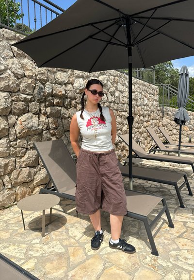 Camisa sin mangas gris claro con un gráfico rojo, pantalones cortos de cargo marrones, zapatillas negras, de pie al lado de una mesa redonda y una gran sombrilla gris.