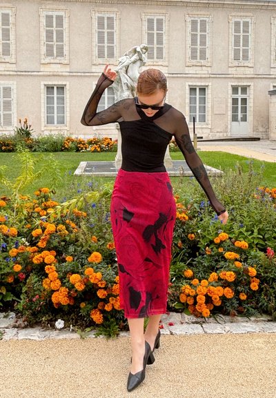 Top negro transparente de manga larga combinado con una falda roja con estampado de rosas y zapatos de tacón negros, frente a un fondo de jardín floral.