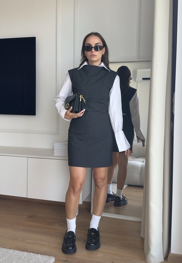 Woman wearing black sleeveless dress over white blouse, black sunglasses, holding black clutch, standing indoors with mirror behind her.