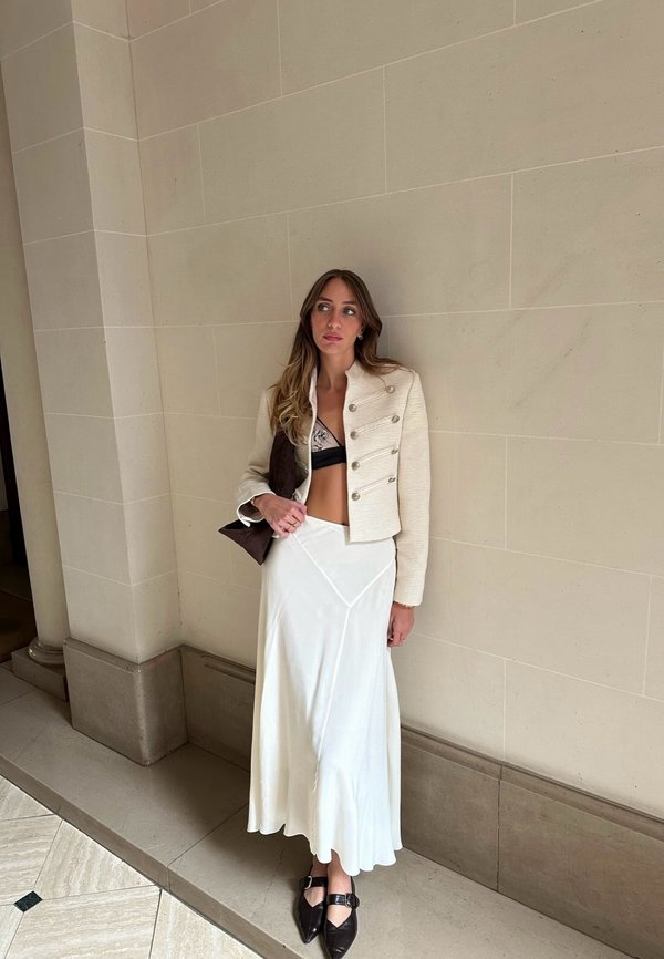 Mujer con chaqueta corta beige, sujetador negro, falda larga blanca y zapatos negros puntiagudos apoyada contra una pared con azulejos en el interior.