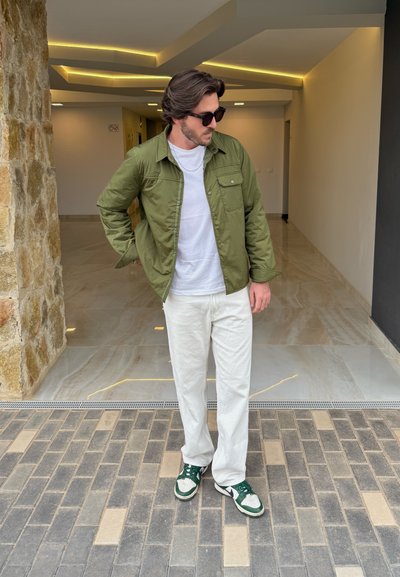 Chaqueta ligera verde oliva sobre una camiseta blanca, acompañada de pantalones blancos y zapatillas verdes. Suelo de ladrillo y piedra en el fondo.