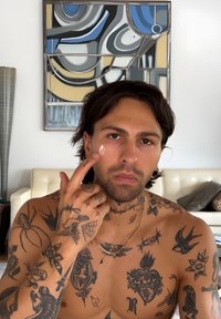 Tattooed man zonder shirt die crème op zijn gezicht aanbrengt, zittend in een moderne woonkamer met een abstract schilderij en een witte bank op de achtergrond.