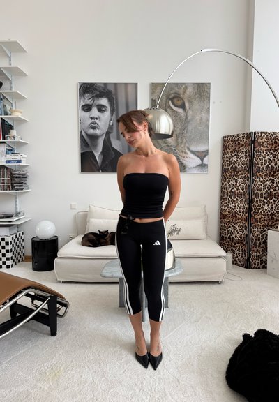 Mujer con top tipo tubo negro y leggings está de pie en una sala moderna con sofá, gato, biombo con estampado de leopardo y grandes fotos de un hombre y un león.