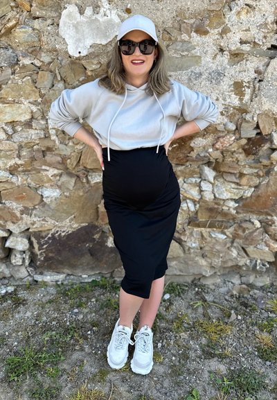 Sudadera gris claro con cordones blancos, falda de maternidad negra ajustada y zapatillas deportivas blancas gruesas; de pie contra una pared de piedra.