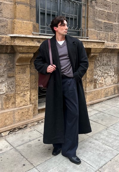 Hombre con abrigo largo negro, suéter gris y pantalones oscuros de pie sobre una acera de piedra, sosteniendo un bolso de hombro color granate junto a una pared de piedra.