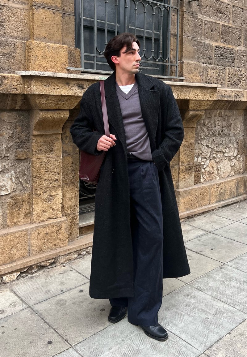 Man in lange zwarte jas, grijze trui en donkere broek staat op een stenen trottoir, met een kastanjebruine schoudertas naast een stenen muur.