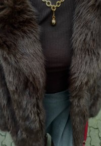 Manteau en fausse fourrure brun foncé avec des fibres longues et douces. Le haut superposé est côtelé, brun foncé, assorti d'une jupe gris clair. Collier chaîne dorée avec un pendentif rond.