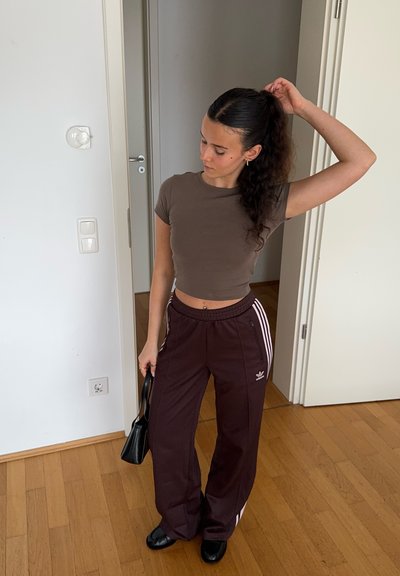 Camiseta corta marrón combinada con pantalones de chándal burdeos con rayas blancas a los lados. Sostiene un pequeño bolso negro. Fondo de suelo de madera.