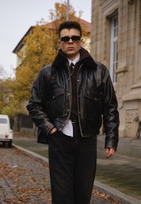 Sort læderjakke med krave af imiteret pels, der har fire forlommer foran og lynlås, kombineret med mørke jeans og en hvidstribet skjorte.