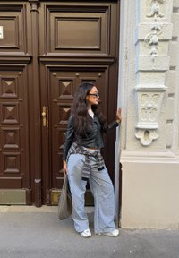 Casaco de couro preto, blusa branca, calças largas listradas, bolsa cinza. Em frente a portas duplas castanhas com painéis e uma parede texturizada branca.