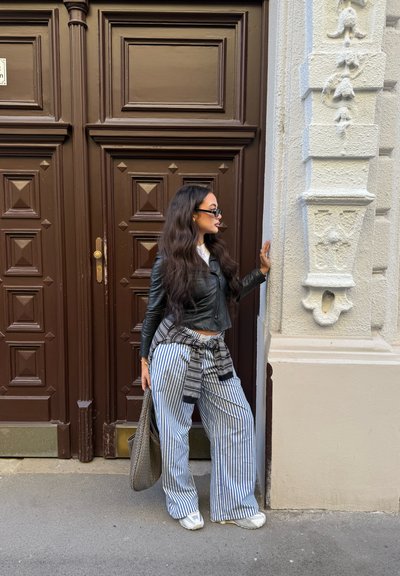 Chaqueta de cuero negra, camiseta blanca, pantalones de pierna ancha a rayas, bolso gris. Frente a puertas dobles marrones con paneles y pared blanca texturizada.