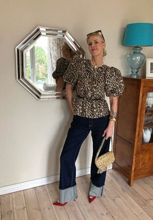 Blusa de mangas abullonadas con estampado de leopardo, vaqueros oscuros con puños de denim, tacones rojos de punta, y un bolso de cuentas dorado. Espejo hexagonal plateado en el fondo.