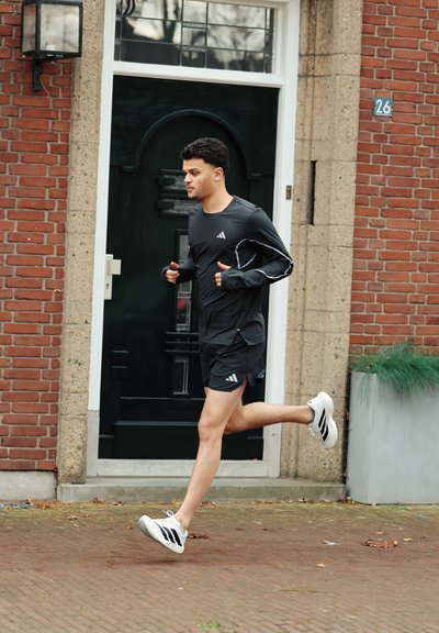 Joven corriendo con ropa deportiva negra y zapatillas blancas sobre una acera de ladrillo frente a una puerta verde y un edificio de ladrillo.