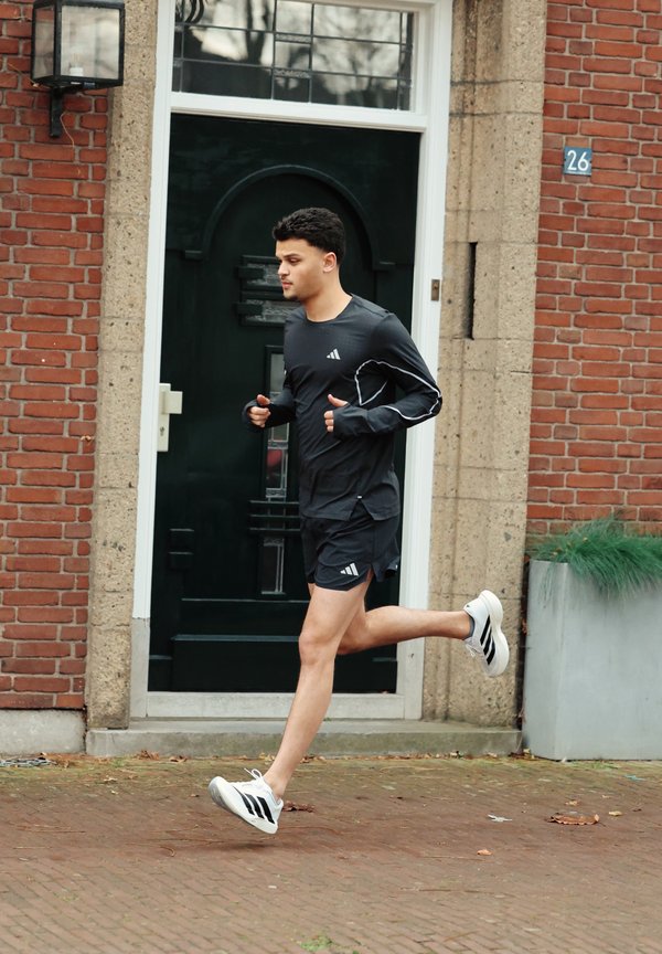 Νέος άνδρας τρέχει φορώντας μαύρα αθλητικά ρούχα και άσπρα αθλητικά παπούτσια σε πεζοδρόμιο από τούβλα μπροστά από μια πράσινη πόρτα και κτίριο από τούβλα.