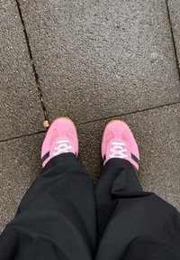 A person wearing pink trainers with white laces and black stripes is standing on wet, textured pavement slabs.