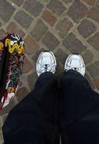 Baskets blanches sur pantalon sombre debout sur un pavé en pierres à côté d'un sac coloré orné de perles.
