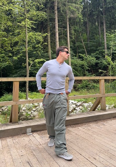 Camisa henley de manga larga gris claro combinada con pantalones cargo de corte holgado en verde oliva, acentuados con cordones naranjas. Zapatillas de cuadros en blanco y negro.