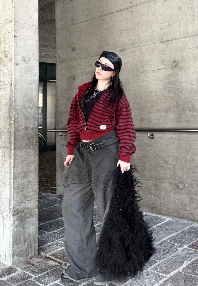 Femme portant des lunettes de soleil noires et un béret en cuir, vêtue d'une veste rayée rouge, d'un pantalon gris ample, et tenant un grand sac noir et duveteux près d'un mur en béton.