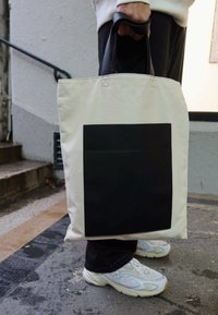 Krém színű vászontáska, nagy fekete műbőrtasakkal. Fekete fogantyúk; téglalap alakú. Földszinti nézet, texturált felület előtt.