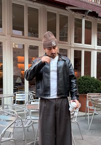 Svart skinnjakke med glidelåslukking, med krage og sidelommer, kombinert med grå vidbukser og en hvit henley-skjorte.