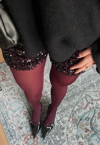 Must värviga ülepaisutatud sviiter, sädelevad buršaani lühikesed püksid, millel on pärlid, buršaani sukkpüksid ja mustad terava ninaga kõrged kontsad, millel on pandla. Mustriga vaipa taust.