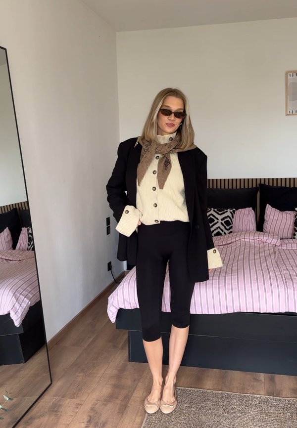 Mujer con gafas de sol, blazer negro, suéter blanco, bufanda marrón, leggings negros y bailarinas beige de pie en un dormitorio con una cama de rayas rosas.