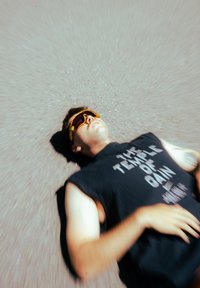 Óculos de sol com armações amarelas e lentes roxas, usados por uma pessoa numa t-shirt preta sem mangas com texto branco, deitada sobre uma superfície texturada.