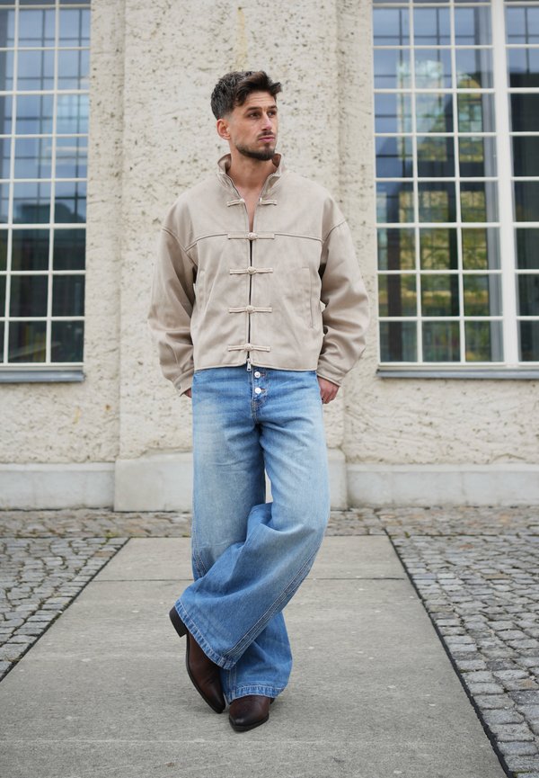Un homme portant une veste beige à brandebourgs et un jean bleu à jambes larges s’appuie contre un bâtiment en pierre doté de grandes fenêtres.