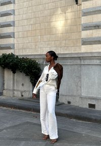 Completino bianco in lino con maniche lunghe e pantaloni a zampa, borsa a tracolla marrone, occhiali da sole e sandali piatti beige, in piedi contro un muro di pietra.