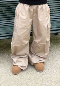 Béžové, nadmerné cargo nohavice z lesklého, textúrovaného materiálu; majú bočné vrecká a elastický pás. Sedia k hnedej semišovej obuvi.