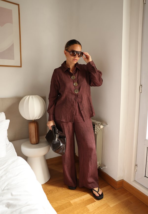 Femme ajustant ses lunettes de soleil portant une chemise marron boutonnée et un pantalon, tenant un sac noir, debout près d'une lampe blanche et d'une fenêtre dans une chambre.
