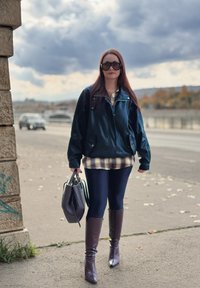 Sort læderjakke, cremefarvet ternet skjorte, marineblå leggings og brune knæhøje støvler. Holder en mørk håndtaske. Bymiljø, overskyet baggrund.