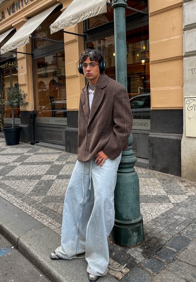 Blazer oversized de cuadros marrones, camiseta blanca, vaqueros baggy azul claro con cremallera, zapatos plateados, de pie junto a un poste verde en un entorno urbano.