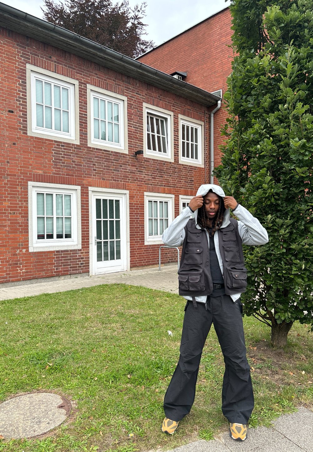 Gilet nero a più tasche indossato sopra a un hoodie grigio, abbinato a pantaloni neri larghi. L'individuo si trova su dell'erba vicino a un edificio in mattoni e a un cespuglio.