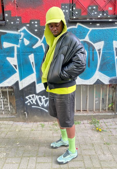 Chaqueta de plumas negra, sudadera con capucha verde neón y gorra, pantalones cortos de cuero negros, calcetines verdes y zapatillas verdes con acentos texturizados, todo contra un fondo de grafitis.