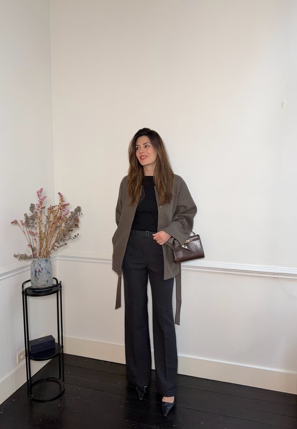 Vrouw die binnen staat en een grijze jas, zwarte top en broek draagt, terwijl ze een bruine handtas vasthoudt, met een vaas gedroogde bloemen op een standaard vlakbij.