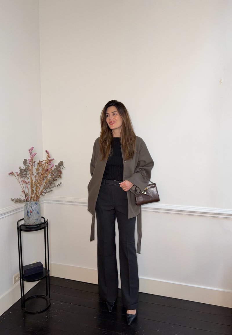 Donna in piedi all'interno che indossa una giacca grigia, top nero e pantaloni, tiene in mano una borsa marrone, con un vaso di fiori secchi su un supporto vicino.