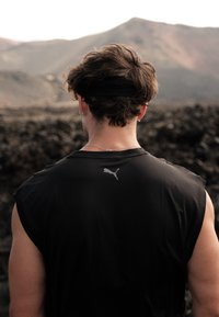 Zwarte mouwloze sportshirt met een zilveren Puma-logo op de achterkant. Heeft een gladde textuur en een ontspannen pasvorm. Lang, golvend haar zichtbaar.