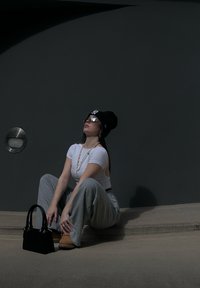 Top corto blanco, pantalones de chándal anchos grises, gorro negro, gafas de sol plateadas, collar de cadena dorada y bolso negro pequeño sobre una superficie de concreto.