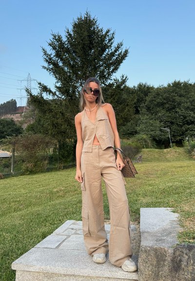 Top corto de lino beige y pantalones de pierna ancha con bolsillos utilitarios. La modelo lleva gafas de sol oscuras y sostiene un bolso de mano estampado. Calzado informal.