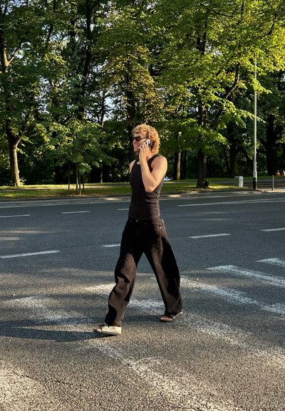 Persona caminando por un camino, vistiendo una camiseta sin mangas negra y pantalones negros holgados, sosteniendo un teléfono, con cabello rizado y gafas de sol. Árboles verdes de fondo.
